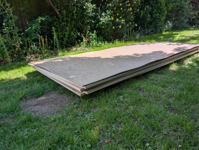 A pile of plywood and hardboard sheets sits in the garden, ready to be cut to size